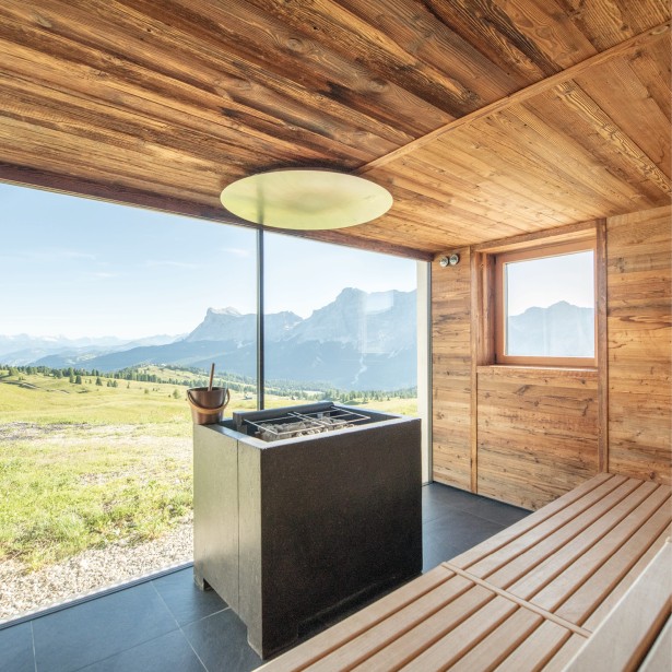 Area sauna con rivestimento in legno e una vista mozzafiato sulle Dolomiti in lontananza. Il secchio per il maestro di sauna e la prossima gettata di vapore è pronto.