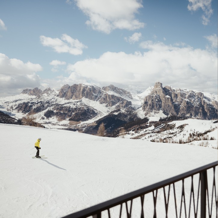Skier in a yellow jacket is skiing on a snow-covered slope with the Dolomites mountain range in the background. In the foreground, a piece of the metal railing from the balcony can be seen.