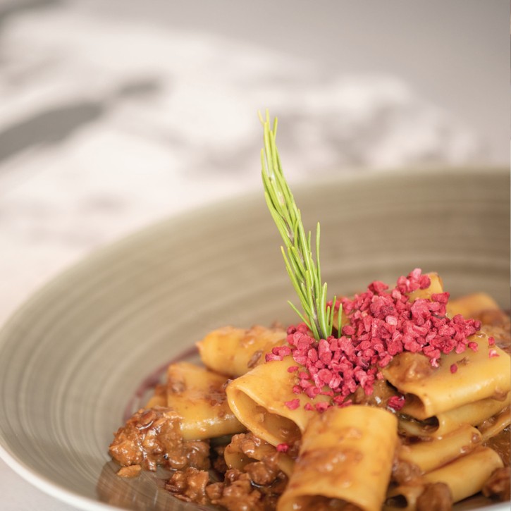 Teller mit Nudeln der Sorte Cannelloni und brauner Bolognese-Soße, dekoriert mit einem Rosmarinstängel und pinken, getrockneten Topping.