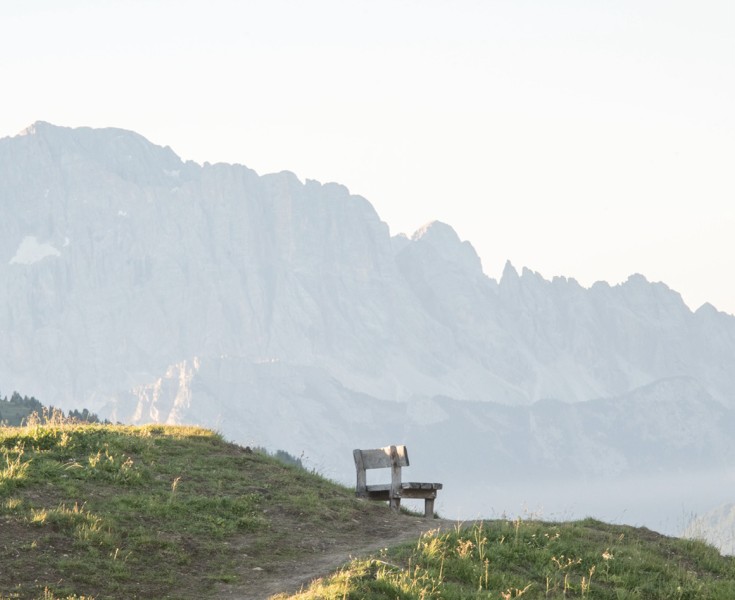 Panchina di legno solitaria su un colle erboso al sole mattutino, con una splendida vista sulle Dolomiti avvolte nella nebbia sullo sfondo.