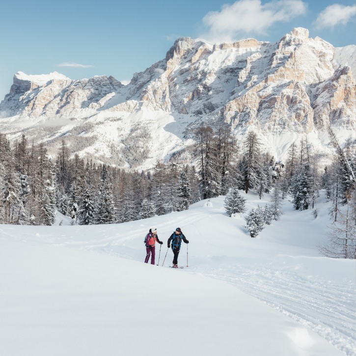 Zwei Personen beim Schneeschuhwandern in der verschneiten Berglandschaft mit Nadelbäumen und markanten Dolomitenfelsen im Hintergrund.