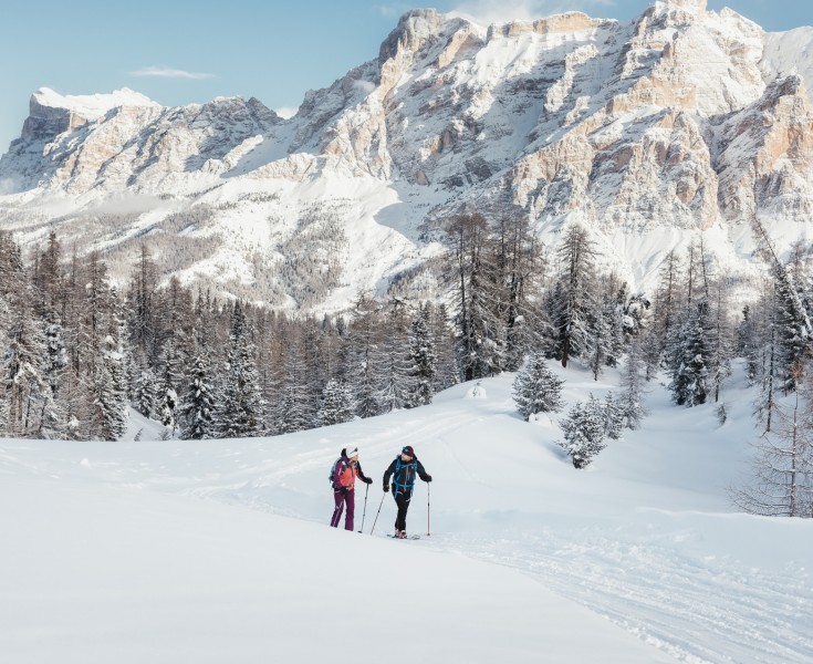 Due persone che fanno escursioni con le ciaspole nel paesaggio montano innevato con conifere e imponenti rocce dolomitiche sullo sfondo.