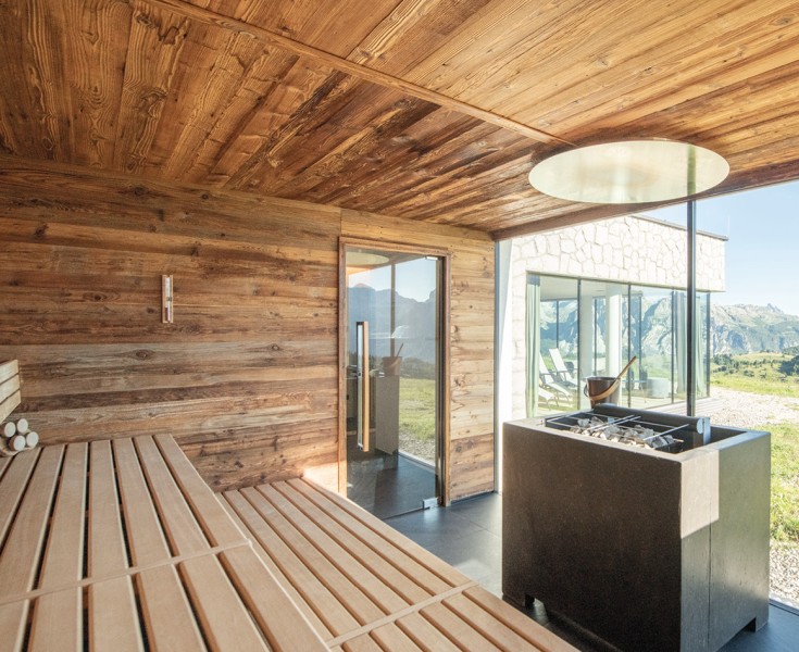 La sauna luminosa, rivestita in legno, con stufa quadrata e ampie vetrate. Vista sul paesaggio montano solitario intorno a Pralongià.