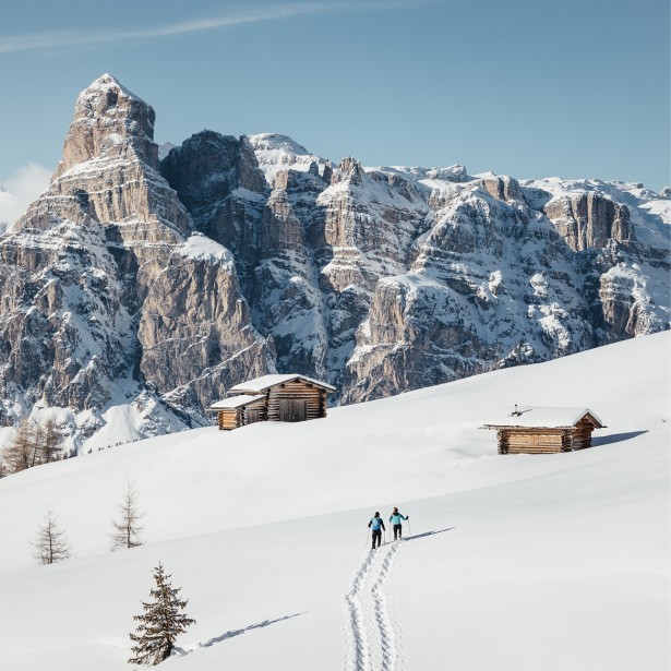 Two people are snowshoeing through the untouched and sun-drenched snowy landscape right below the impressive Sassongher, a well-known Dolomite mountain. They are the only ones leaving footprints.