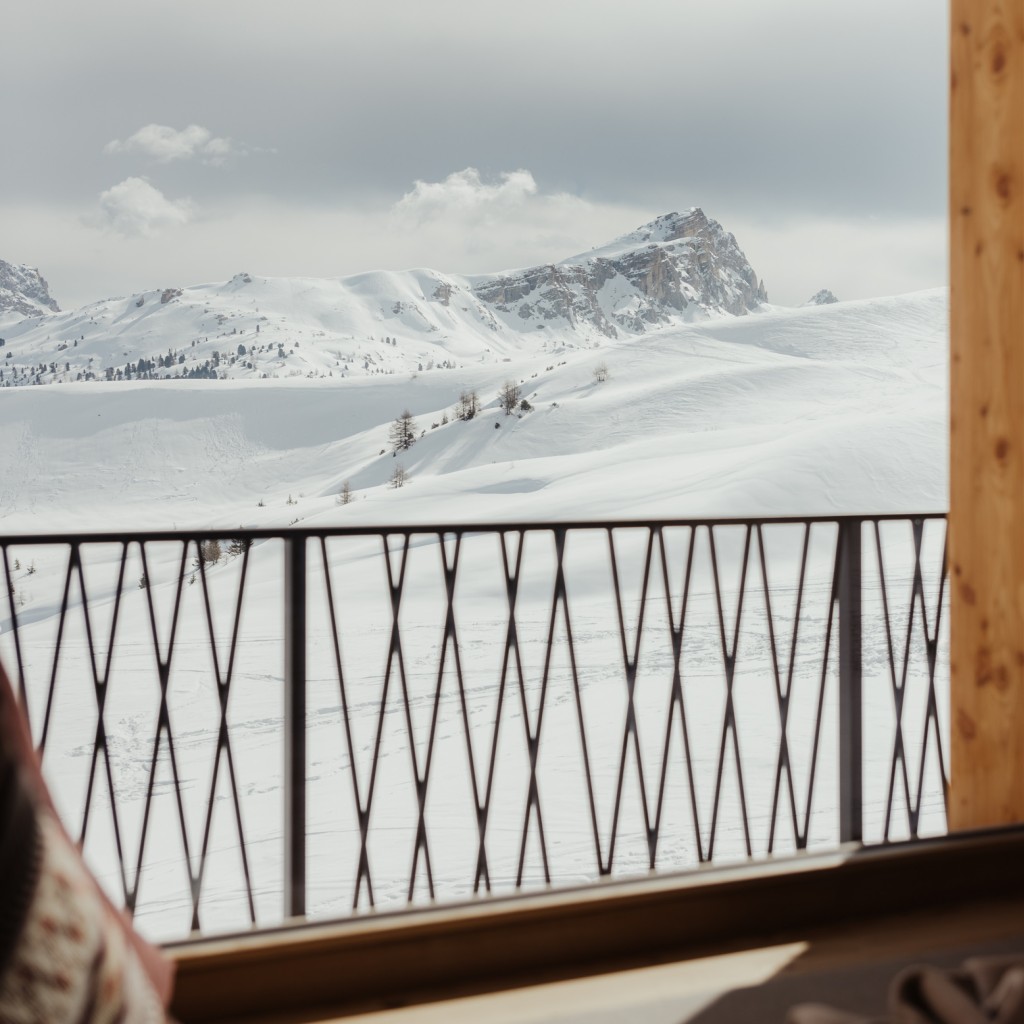 Snow-covered mountain landscape with isolated trees, a Dolomite peak in the background, and a metal balcony railing in the foreground.