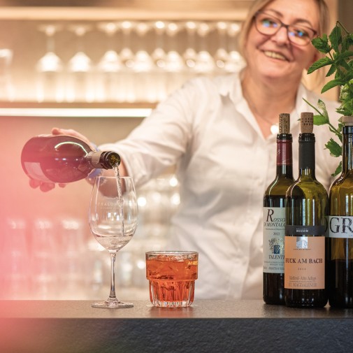 La cameriera del Rifugio Alpino Pralongià sta versando un bicchiere di vino bianco ridendo. Accanto, un bicchiere con una bevanda rossa brillante e altre tre bottiglie di vino si trovano sul bancone.