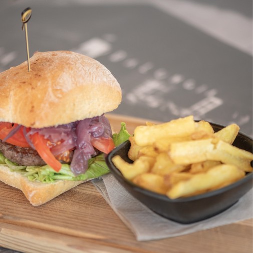 Sandwich with onions, tomatoes, lettuce, and meat, served with a bowl of fries on a wooden board.