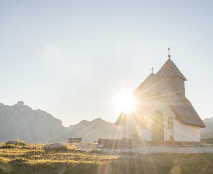 Piccola cappella circondata da un prato e illuminata dal sole. Lo splendido panorama montano è visibile sullo sfondo.