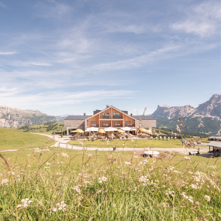 The Pralongià mountain inn in a wonderful solitary location, with sun umbrellas on the terrace, surrounded by a blooming, summer flower meadow. Mountains and sky in the background.