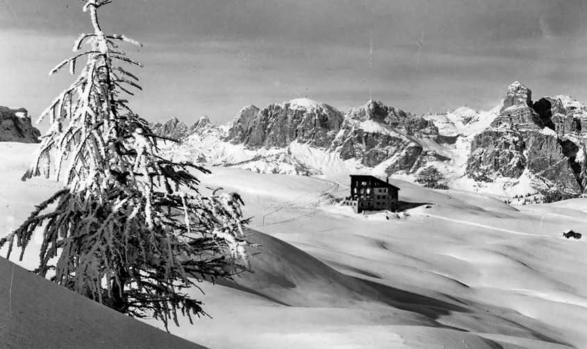 Old black-and-white photo with the old Pralongià mountain inn, standing solitary in the midst of the snow-covered mountain landscape.
