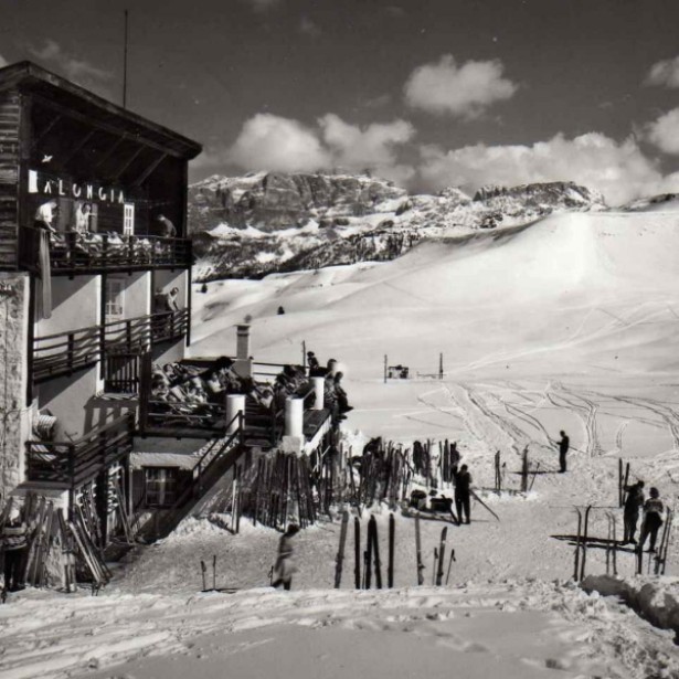 Old black-and-white photo of the Pralongià mountain inn in winter. Next to it are the snow-covered ski slope and the mountains in the background.